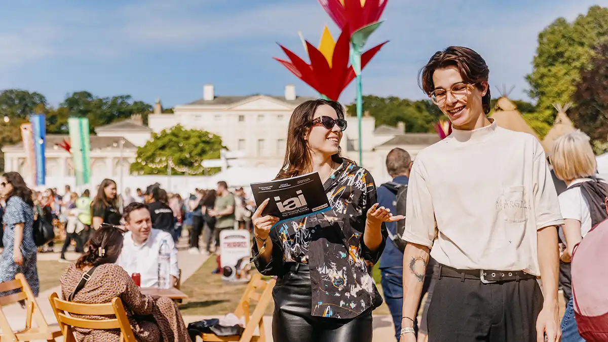  A lively outdoor festival scene featuring a woman holding a program and talking to a casually dressed man, surrounded by colorful decorations and attendees. 