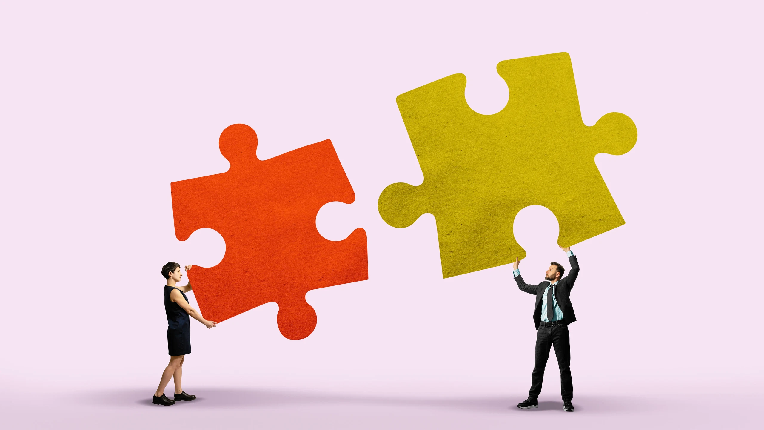  A man and a woman hold large puzzle pieces, one red and one yellow, symbolizing teamwork and connection against a light pink background. 