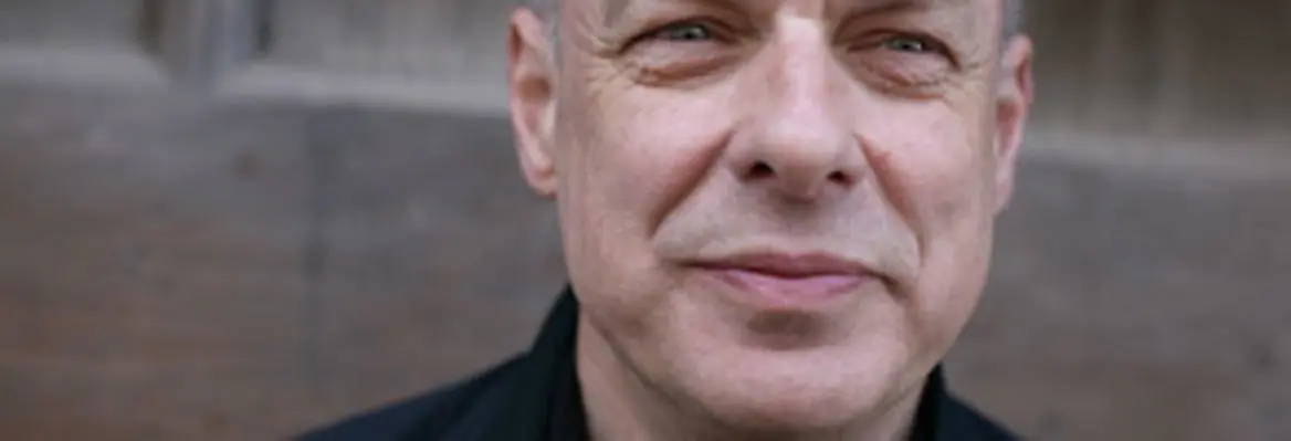A close-up of a middle-aged man with gray hair, smiling slightly, wearing a black jacket against a wooden background.