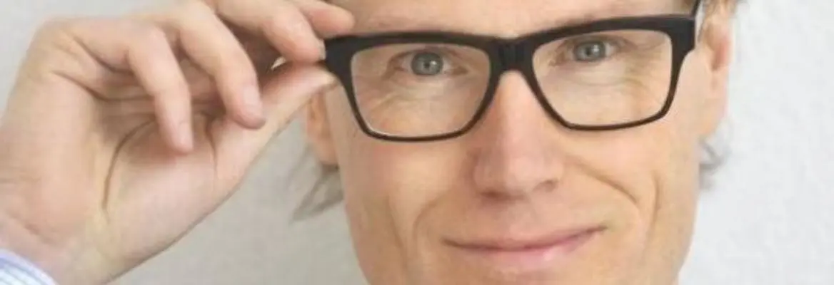 A smiling man with glasses adjusts them, showcasing a professional look with a slight smirk against a neutral background.