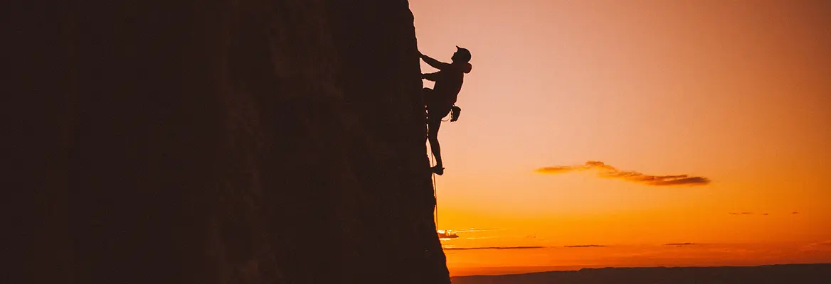 A lone rock climber grips a steep cliff in silhouette against a glowing orange sunset, creating a dramatic, adventurous scene of courage and solitude.