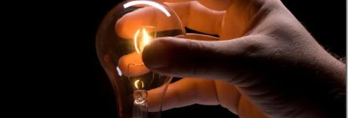 A person's hand holding a light bulb, with a glowing filament inside, set against a dark background.