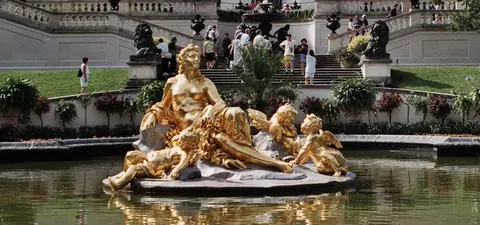 A golden statue of a woman with children sits in a pond, surrounded by lush greenery and people strolling in the background.