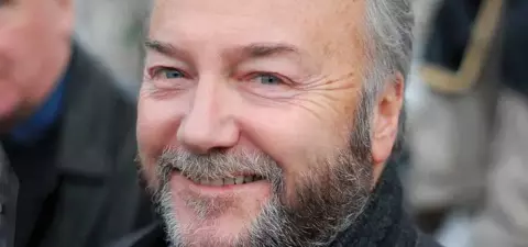 A smiling man with a beard, wearing a dark coat, stands outdoors with blurred figures in the background.