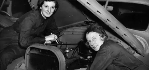 Two women in overalls smiling while working on a car engine, with the hood raised.