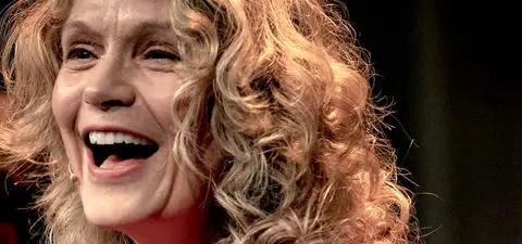 Smiling woman with curly hair, captured during a performance or event, radiating joy and enthusiasm. Soft lighting highlights her expression.