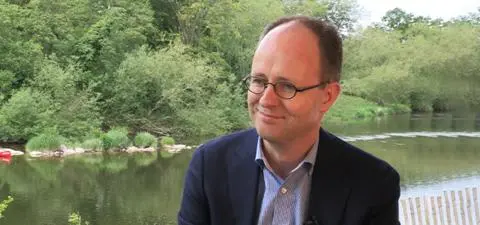 A smiling man with glasses sits outdoors near a river, surrounded by lush greenery, wearing a blue suit and a light-colored shirt.