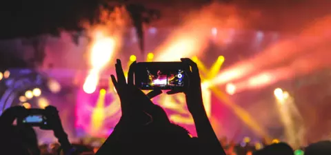 A crowd at a concert, with hands holding smartphones capturing the performance, surrounded by vibrant stage lights and a lively atmosphere.
