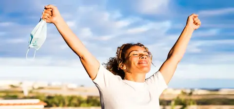 A woman with curly hair joyfully raises her arms in the air, holding a face mask, against a bright sky and scenic backdrop.