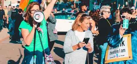 A diverse group of activists participates in a colorful march, with one woman on a megaphone and another using a smartphone, holding a sign advocating for rights.