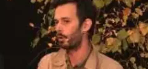 A man with a beard speaks in front of a backdrop of green leaves, wearing a light-colored jacket.