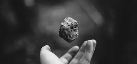 A black and white image of a hand reaching out to catch a small rock floating above it, surrounded by a blurred natural background.