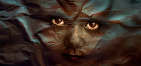 A close-up of a child's face partially obscured by textured, crumpled layers, creating a mysterious and surreal effect with deep, expressive eyes.