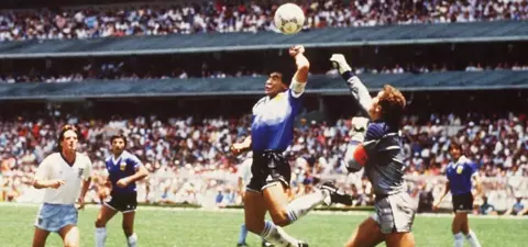 Diego Maradona leaps to make a crucial play during a football match, with players in action and a packed stadium in the background.