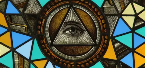 Stained glass window featuring a prominent Eye of Providence within a triangle, surrounded by colorful geometric shapes.