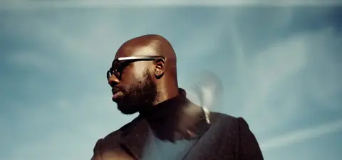 A man with a bald head and glasses stands against a blue sky, wearing a dark coat and a turtleneck.