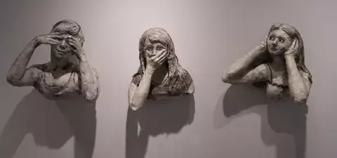 Three abstract sculptures of women with varying expressions, each covering different parts of their faces, set against a plain wall.