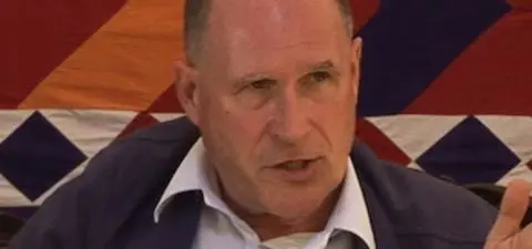 A man in a blue jacket gestures while speaking, with a colorful geometric fabric backdrop behind him.