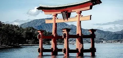 A large, traditional Japanese torii gate stands in the water against a backdrop of mountains and a clear sky.