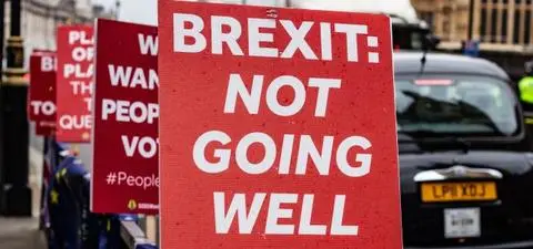 Protest signs display messages about Brexit, highlighting public dissatisfaction with the process, in a city setting with vehicles in the background.