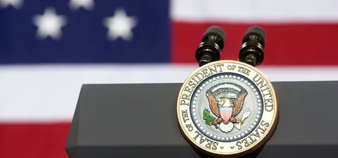 A podium featuring the presidential seal of the United States, set against a backdrop of the American flag with stars and stripes.