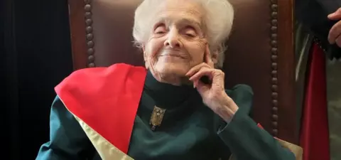 An elderly woman with white hair sits comfortably in a chair, smiling slightly while dressed in formal attire with a distinctive sash.
