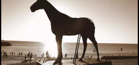 A silhouetted large horse sculpture stands against the sunset, overlooking a beach where people are enjoying the scenery.