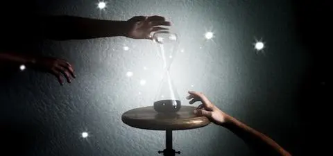 An hourglass glows in the dark, surrounded by shimmering lights, as two hands reach towards it, symbolizing the passage of time.