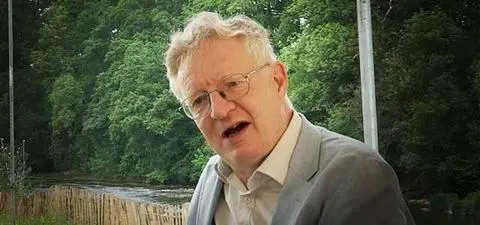 A middle-aged man with glasses in a light suit, speaking thoughtfully against a backdrop of lush green trees and a stream.