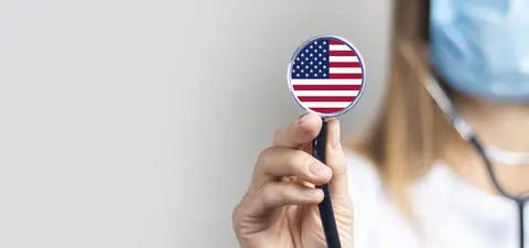 A healthcare professional wearing a mask holds a stethoscope with an American flag design.