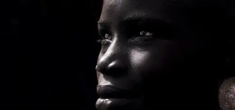 A close-up portrait of a woman with dark skin, featuring a serene expression and prominent earrings, against a dark background.
