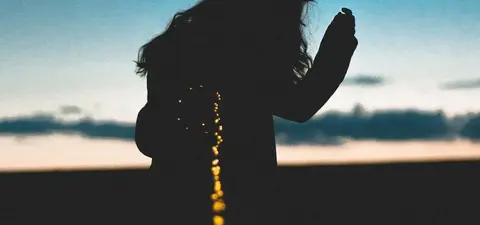 Silhouette of a person with wavy hair against a sunset sky, holding an object, with a glowing trail extending behind them.