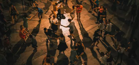 A vibrant crowd dances in a lively open space, with diverse groups enjoying the music and rhythm, creating dynamic shadows on the ground.
