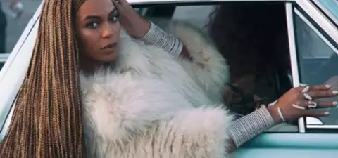 A woman with long braided hair and a luxurious fur coat poses in the window of a classic car, showcasing an elegant style.