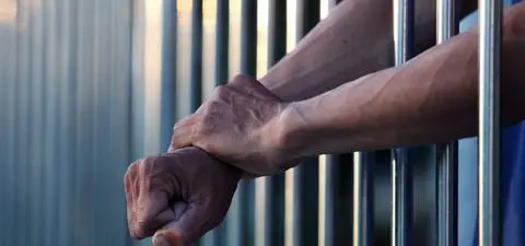 A pair of hands gripping prison bars, one hand clenched into a fist, symbolizing confinement and struggle.
