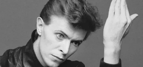 A monochrome portrait of a young man with slicked-back hair and a leather jacket, striking a contemplative pose with one hand raised.