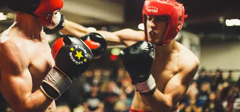 Two male boxers in a ring, wearing headgear and gloves, engaged in a competitive match, with an audience visible in the background.
