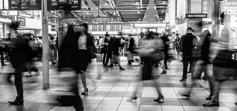 A busy train station in black and white, featuring blurred figures of commuters moving quickly across the tiled floor, with signs in Japanese and English.