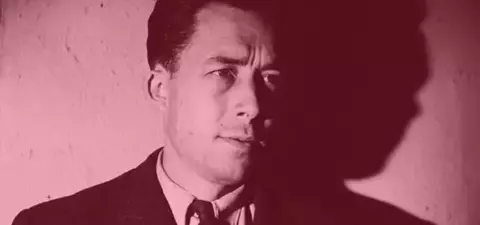 A close-up portrait of a man in a suit with a serious expression, casting a shadow on a textured wall, illuminated in a warm tone.