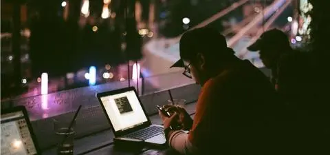 A person sitting at a wooden table, focused on their phone, surrounded by laptops and illuminated city lights in the background.