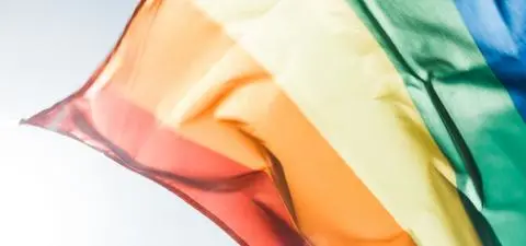 A close-up of a waving rainbow flag, representing LGBTQ+ pride, with vibrant colors including red, orange, yellow, green, blue, and purple.