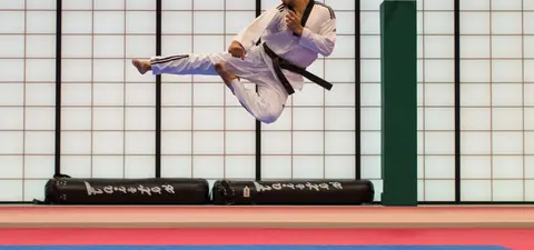 A martial artist in a white uniform performs a high kick in mid-air against a backdrop of a colored training facility.