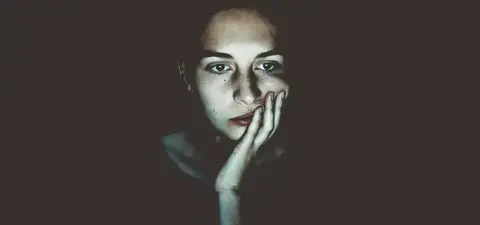 A close-up of a thoughtful young woman with fair skin and dark hair, illuminated against a dark background, showing an expression of contemplation.