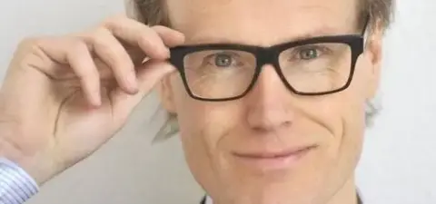 A smiling man with glasses adjusts them, showcasing a professional look with a slight smirk against a neutral background.