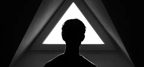 A silhouette of a person standing in front of a triangular window, with stark contrast highlighting the geometric shapes and the figure's outline.