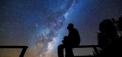 A silhouette of a person sitting on a railing, gazing at a starry sky filled with the Milky Way galaxy and numerous stars.