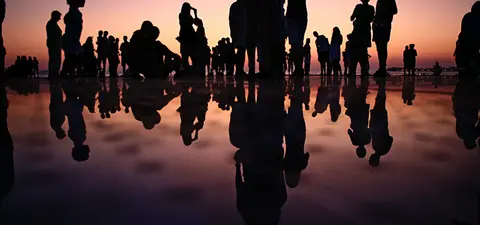 Silhouettes of people gathered at sunset, reflecting in water, with vibrant colors of the horizon creating a serene atmosphere.