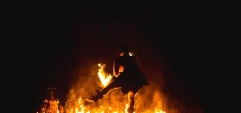 A performer dances with fire amidst flames, creating an intense, dramatic scene against a dark backdrop.