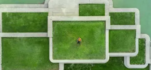 An aerial view of a park featuring geometric stone pathways and patches of green grass, with a person standing in the center.