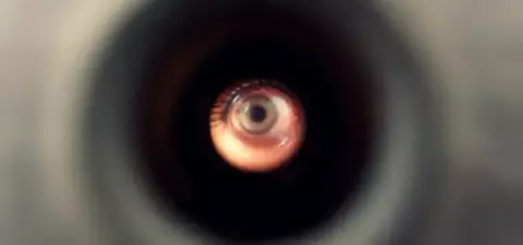 A close-up view of a circular hole, revealing a human eye looking through it, with a blurred background.